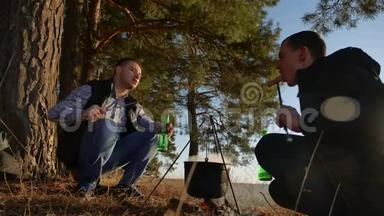 两名游客啤酒正坐在篝火徒步聚会上，在大自然中露营剪影，阳光日落。 两个剪影